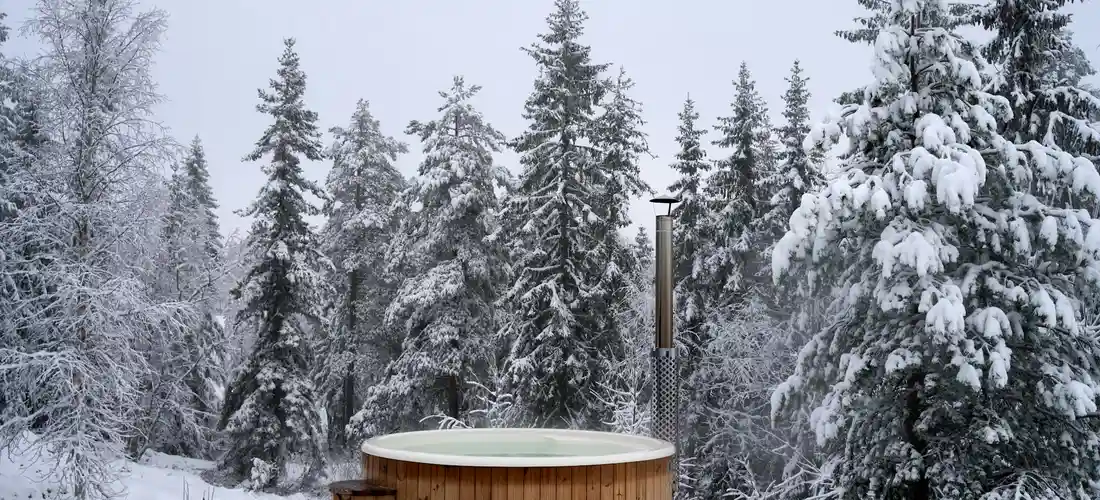 A wooden round hot tub sits outdoors in a snowy forest with tall, snow-covered evergreen trees surrounding it.