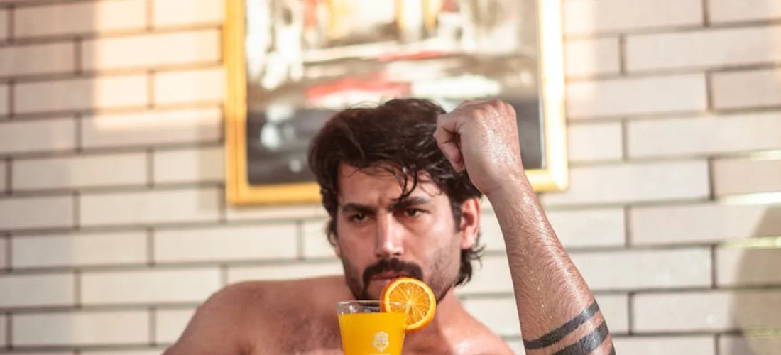 A shirtless man with a tattoo on his forearm sits indoors, holding a glass of orange juice with an orange slice on the rim, in front of a brick wall.