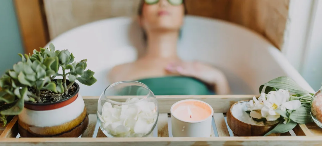 A person relaxing in a spa tub with a wooden tray holding plants, a candle, and a glass of water, highlighting a calm spa setting.