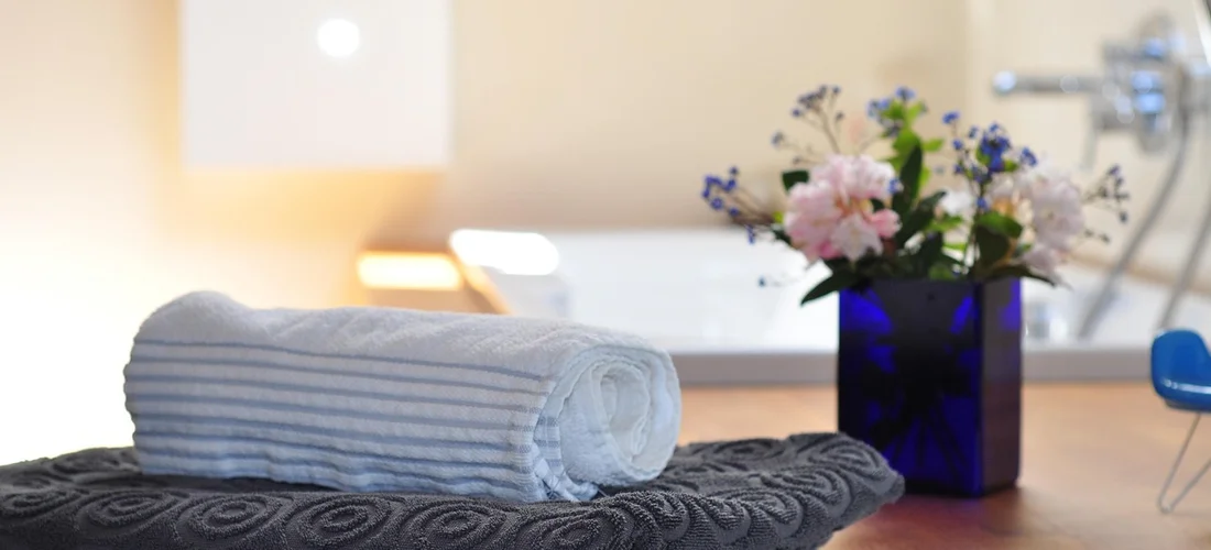 Rolled white towel on a dark patterned spa mat with a blue vase of pink and white flowers on a wooden surface in a calm bathroom/spa setting.