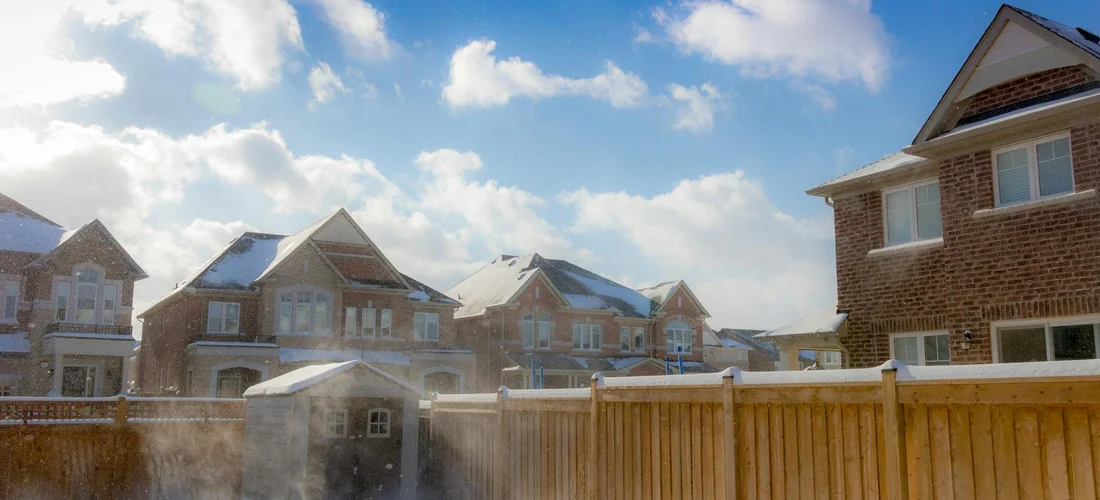 Suburban backyard with a tall wooden privacy fence and brick houses in the background under a blue sky with clouds.