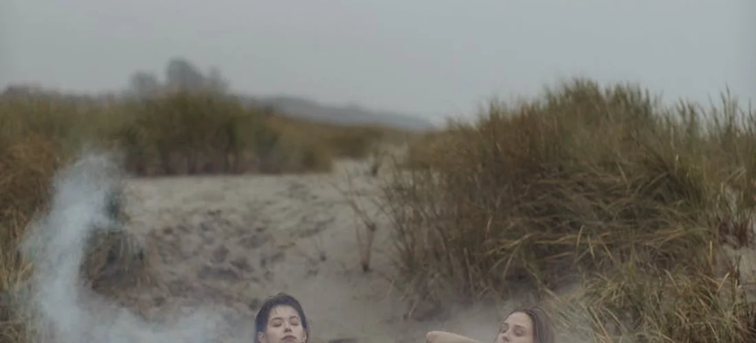 Two people soak in an outdoor hot tub near sandy dunes with patches of grass, steam rising from the water.