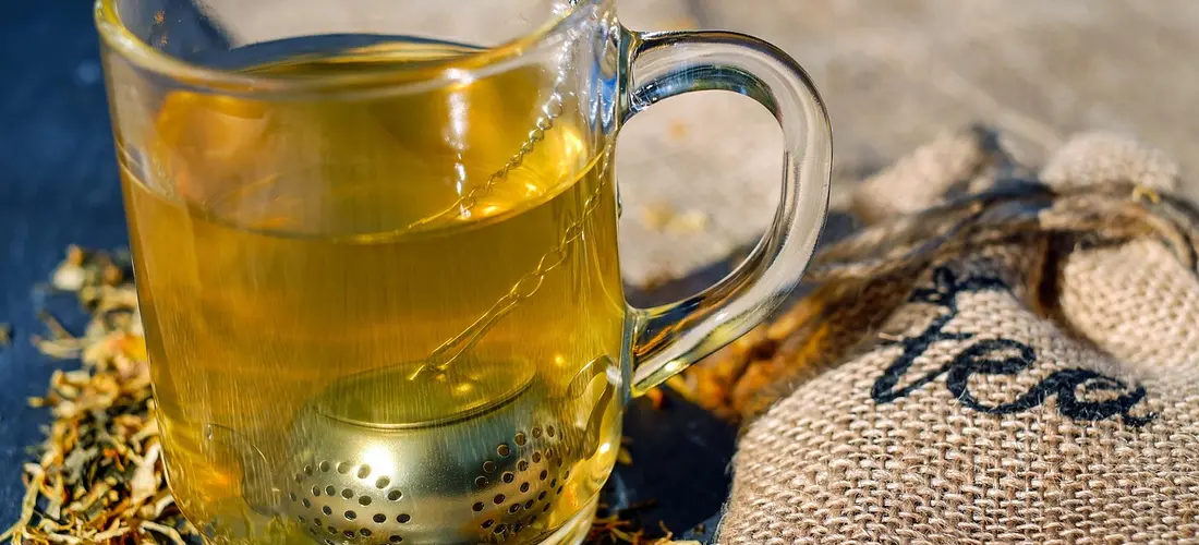 Glass mug of tea with a stainless steel infuser on a rustic surface beside a burlap tea bag.