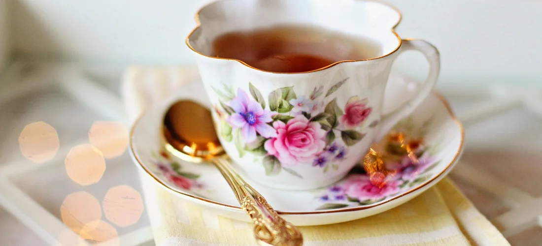 A porcelain teacup with pink and purple floral pattern on a matching saucer, gold spoon beside it, with a soft, warm bokeh background.