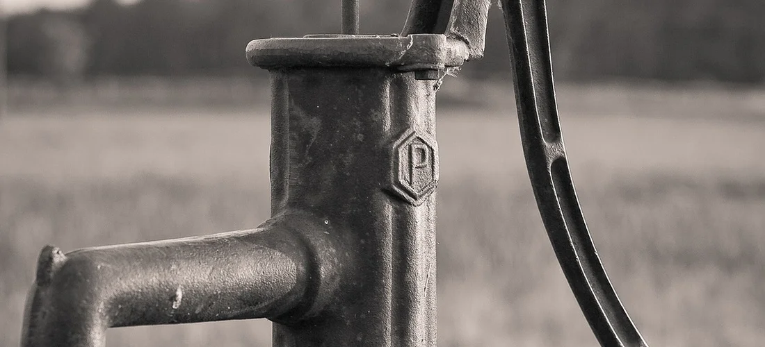 Close-up of a metal water pump with a curved handle in a field.