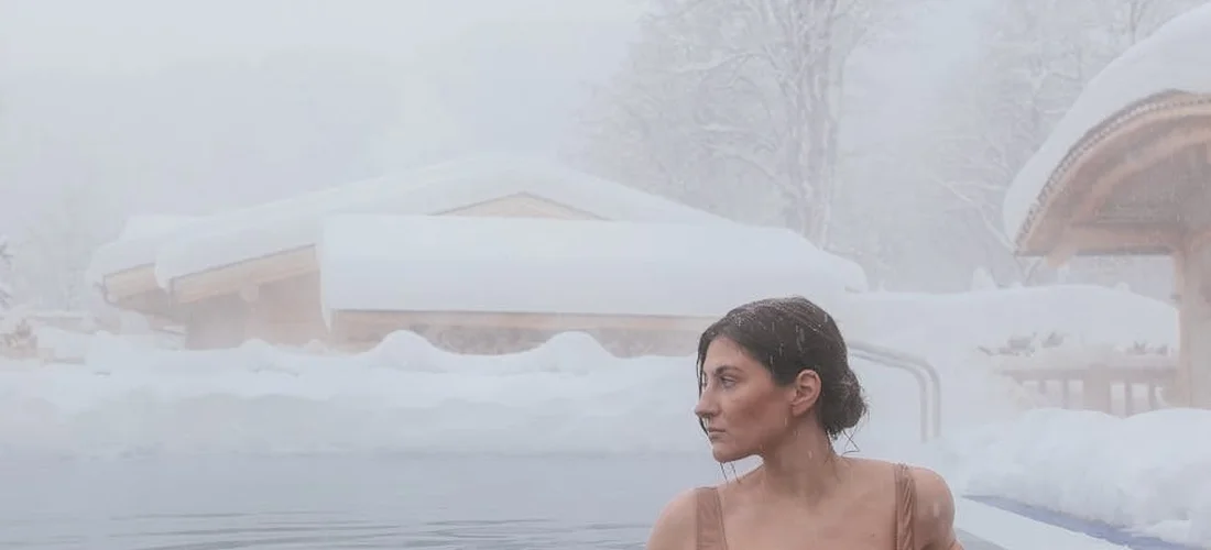 Person relaxing in a hot tub surrounded by a snowy, wintry landscape with snow-covered roofs in the background.