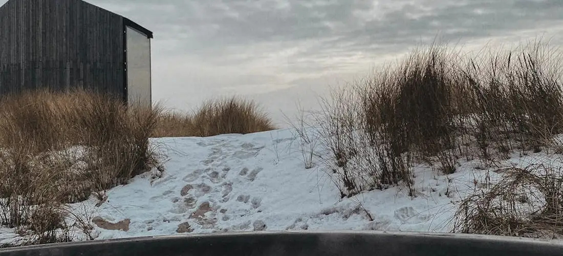 Snow-covered path leading to a dark shed, with dry grasses on the sides and footprints in the snow, viewed from inside a vehicle.