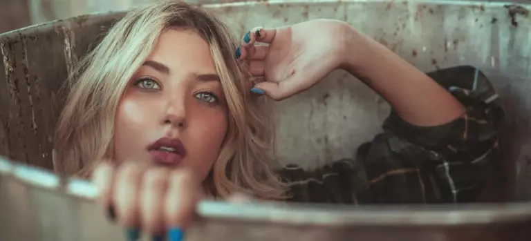 A woman with blonde hair leaning over the edge of a bathtub, looking at the camera.