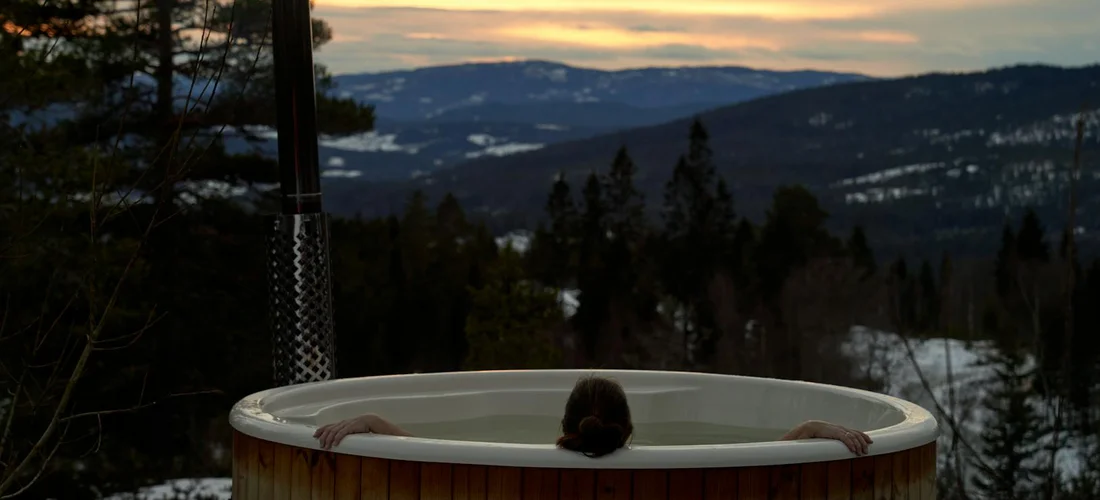 A person soaking in a wooden outdoor hot tub at sunset with pine trees and distant mountains in the background.